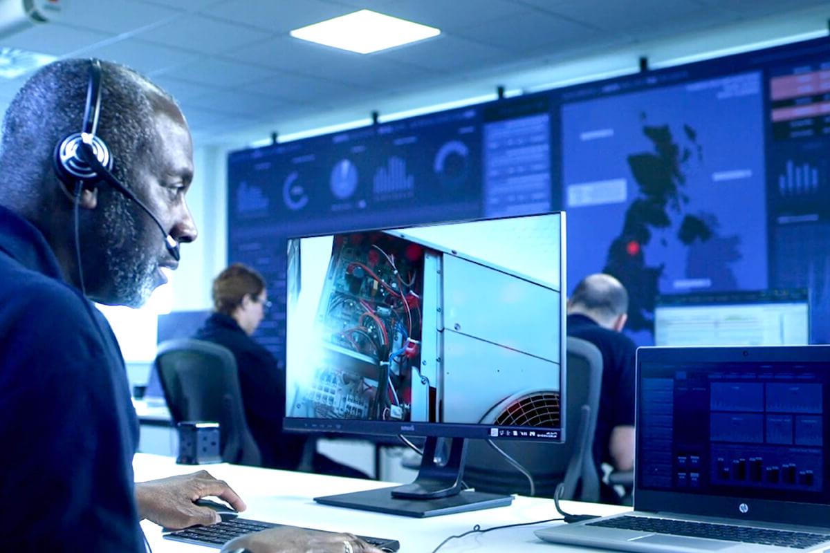 An engineer wearing a headset at a computer terminal, watching maintenance taking place elsewhere. A wall of computer displays 和 other people at desks are in the background