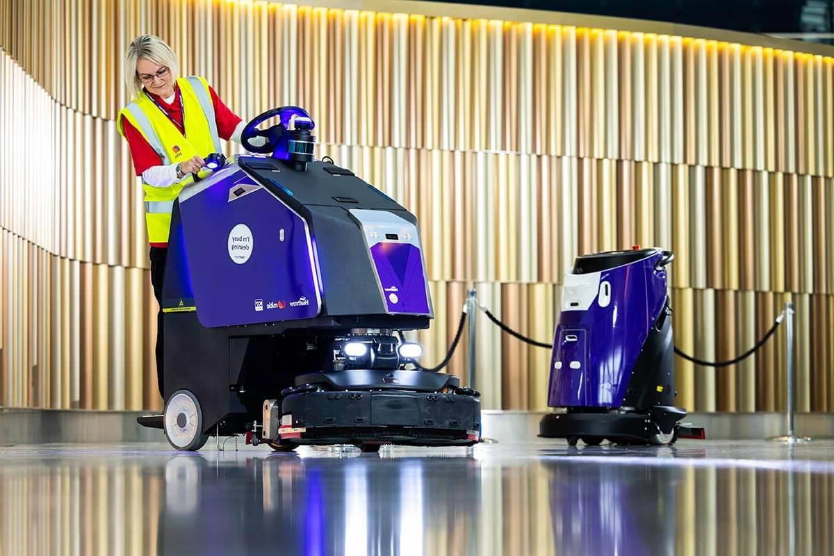 Two purple 和 black Mitie 清洁 robots in London Heathrow Airport, with a female Mitie worker in a high vis vest using the right-h和 robot